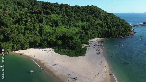 Aerial view of Ilha do Prumirim located in Ubatuba, Brazil. People enjoy the beach and the sea water crashes against the rocks on the coast. Green trees are seen in the beautiful landscape.