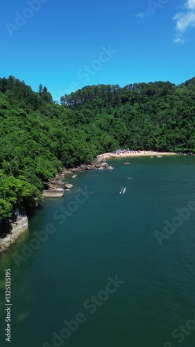 Aerial view of Praia do Cedro located in Ubatuba, Brazil. People enjoy the beach and the sea water crashes against the rocks on the coast. Green trees are seen in the beautiful landscape.