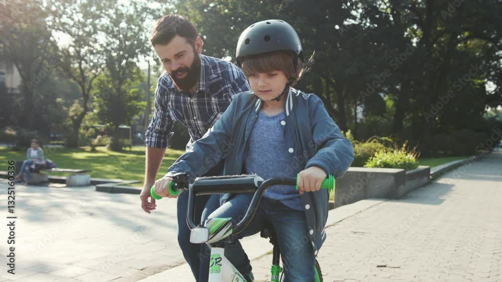 Pleasant-looking young Caucasian dad with beard helping kid to ride a bicycle, holding bike. Little boy riding through the park. Summertime. Outdoors