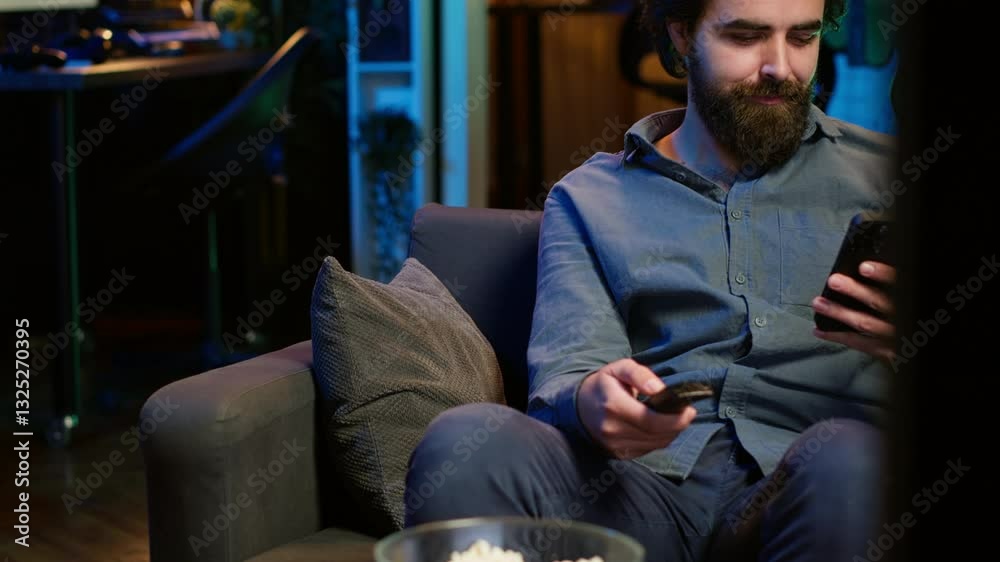 Smiling person on couch picking movie on streaming service using remote and texting on mobile phone. Happy man watching shows on television, zapping though channels, chatting smartphone, camera A
