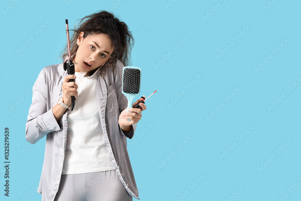 Hurrying African-American woman in pajamas with curling iron, brush and lipgloss talking by mobile phone on blue background Hurrying African-American woman in pajamas with curling iron, brush and lipgloss talking by mobile phone on blue background