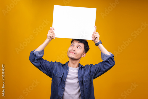 Studio Portrait Of Young Asian Man Holding Up A Blank Mockup Paper Banner Over Yellow Background