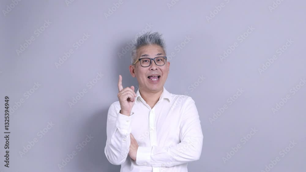 An East Asian senior businessman in a white shirt with glasses and grey hair is standing against a grey background. An East Asian senior businessman in a white shirt with glasses and grey hair is standing against a grey background.