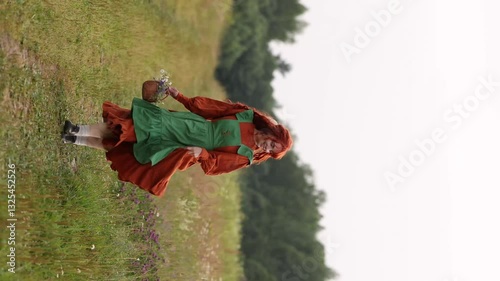 A young red-haired woman in a red and green vintage dress runs along a rural path, looking back cheerfully. Romantic rural scene, the epitome of freedom or rural life.