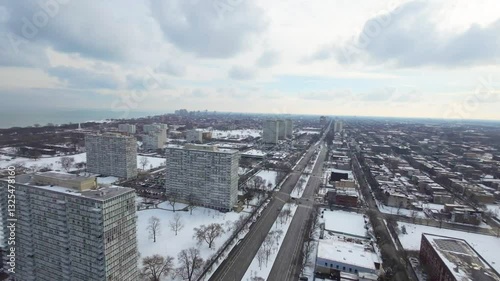 Aerial winter view of a sprawling cityscape with snow-covered trees and rooftops. High-rise buildings roads, parked cars, and distant neighborhoods stretch toward the horizon beneath a partly cloud