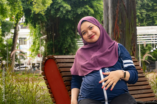 a pregnant Asian Muslim woman, sitting relaxing in a park while looking at her handphone
