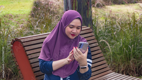 a pregnant Asian Muslim woman, sitting relaxing in a park while looking at her handphone