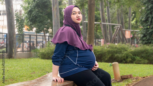 an Asian Muslim woman who is pregnant, is in a green park full of trees, to reduce the stress of facing childbirth