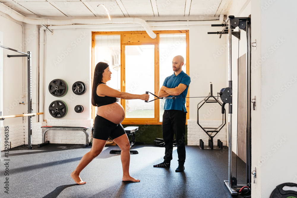 Fototapeta premium Pregnant Chinese Woman Exercising With Resistance Cables Under the Guidance of a Trainer in a Gym Setting