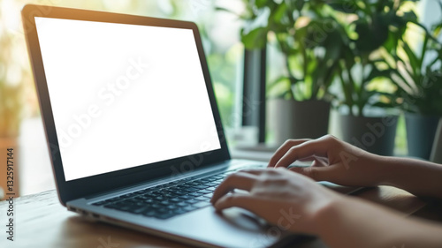 A person working on a laptop with a blank white screen in a bright workspace filled with green plants and natural light.