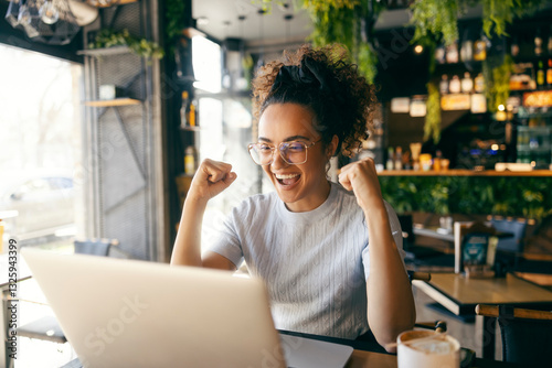 Cheerful female entrepreneur sitting in cafe, looking at laptop and cheering for winning price. photo