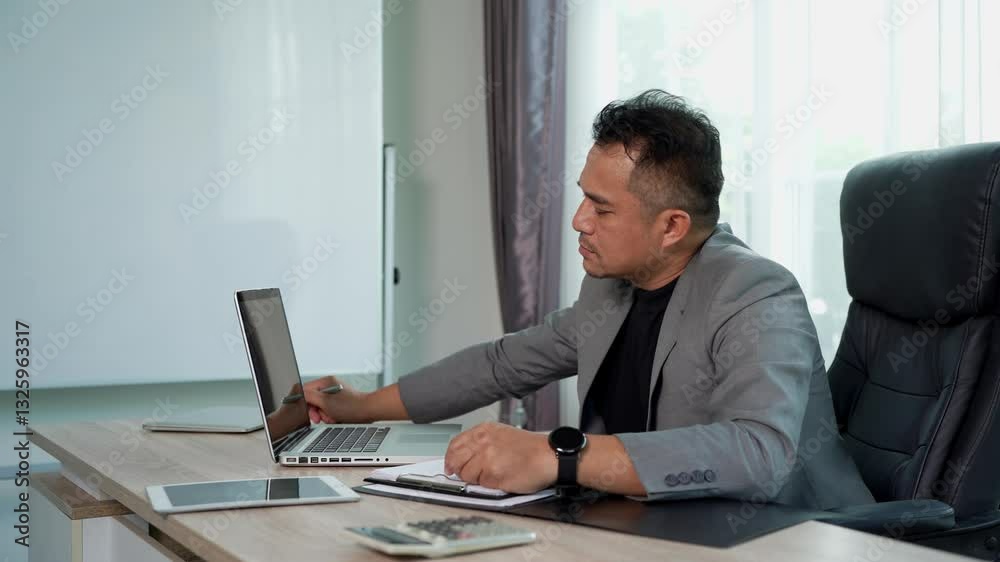 Professional businessman holding a pen in hand, closed his eyes and thoughtful during working on laptop computer, He concentrate his work, various Technology Device placed on desk in modern office Professional businessman holding a pen in hand, closed his eyes and thoughtful during working on laptop computer, He concentrate his work, various Technology Device placed on desk in modern office