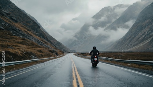 A lone motorcyclist travels along a damp, empty road surrounded by steep, mist-covered mountains during cloudy weather. The atmosphere is tranquil and serene
