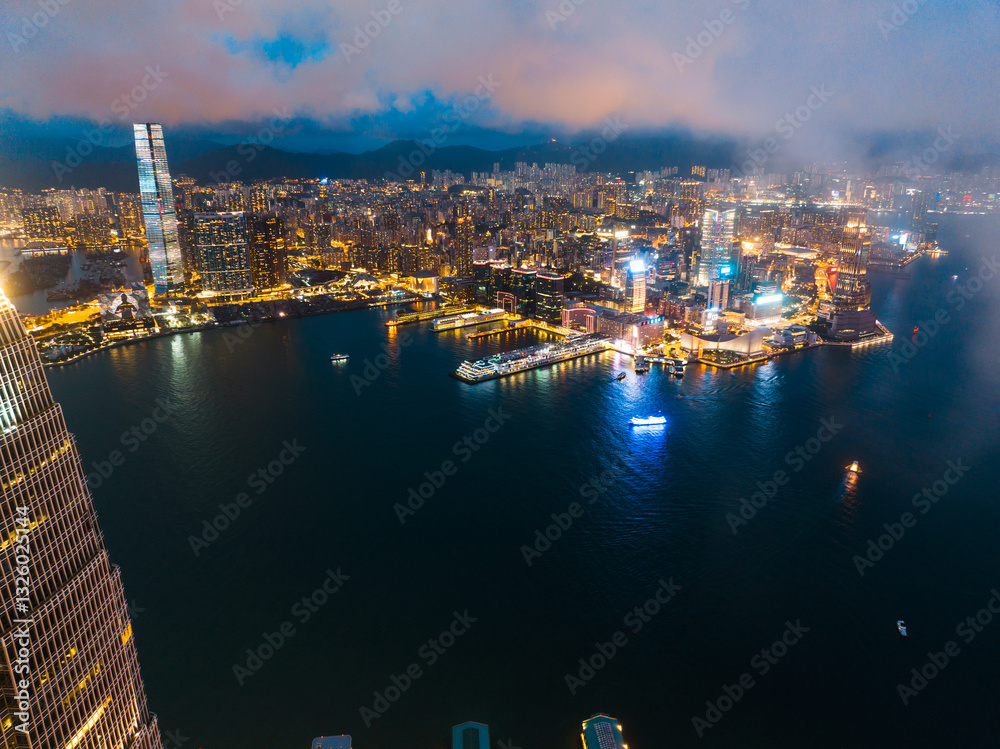 Obraz premium Aerial panorama of Hong Kong city at night, showcasing the skyline, water, and lights under a cloudy sky.