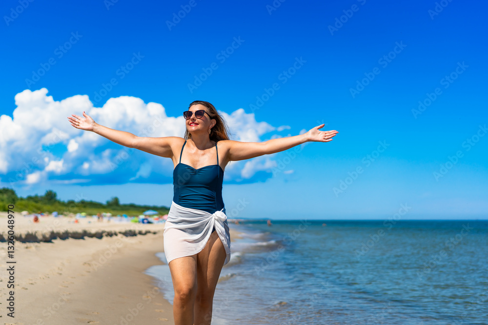 Beautiful middle-aged woman in swimsuit walking with outstretched arms on sandy beach in summertime. Energy and happiness. Front view Beautiful middle-aged woman in swimsuit walking with outstretched arms on sandy beach in summertime. Energy and happiness. Front view