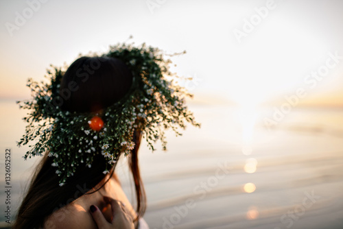 A girl in a wreath of wildflowers stands by the sea at sunset
