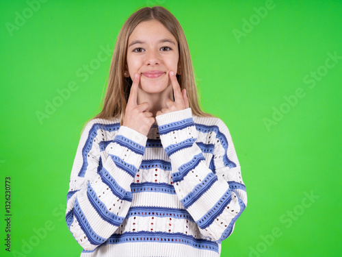 Happy beautiful caucasian girl wearing striped sweater with toothy smile, keeps index fingers near mouth, fingers pointing and forcing cheerful smile