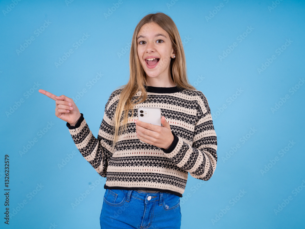 Obraz premium Astonished beautiful caucasian girl wearing striped sweater holding her telephone and pointing with finger aside at empty copy space