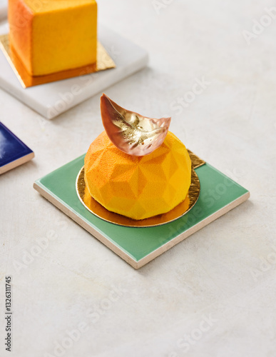 Delicate desserts are artfully arranged on colorful plates in a pastry shop. The bright yellow treat resembles a citrus fruit, topped with metallic leaf for added flair photo