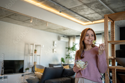 Happy woman eating fresh salad in modern apartment living room