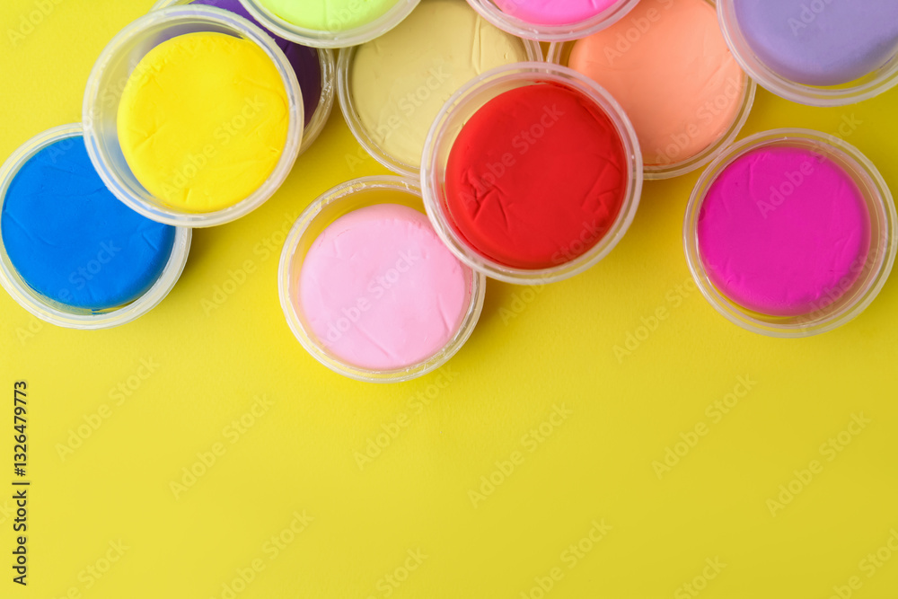 Different colorful modeling clay in plastic jars on yellow background, flat lay. Space for text Different colorful modeling clay in plastic jars on yellow background, flat lay. Space for text