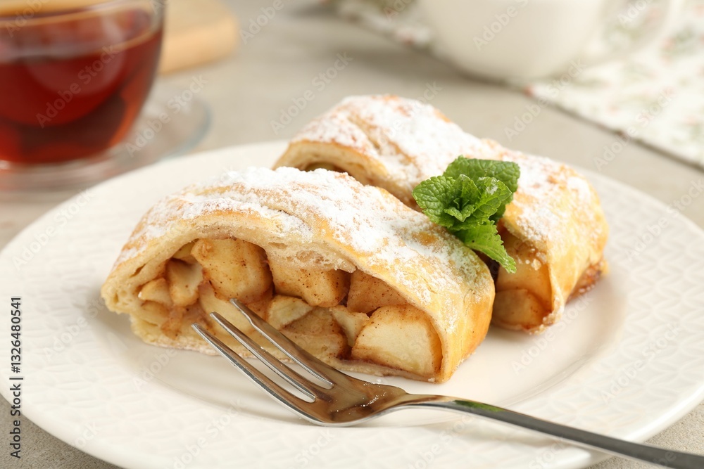 Pieces of tasty apple strudel with powdered sugar, mint and fork on grey textured table, closeup Pieces of tasty apple strudel with powdered sugar, mint and fork on grey textured table, closeup
