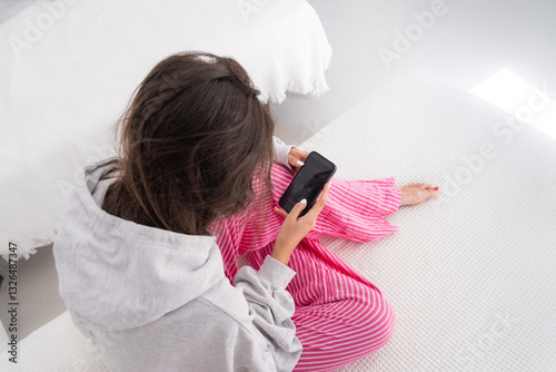 A young girl from behind is sitting on the bed in a white room looks at her smartphone. She is wearing pink pants and a gray sweatshirt and is barefoot. Concept of digital lifestyle and social media