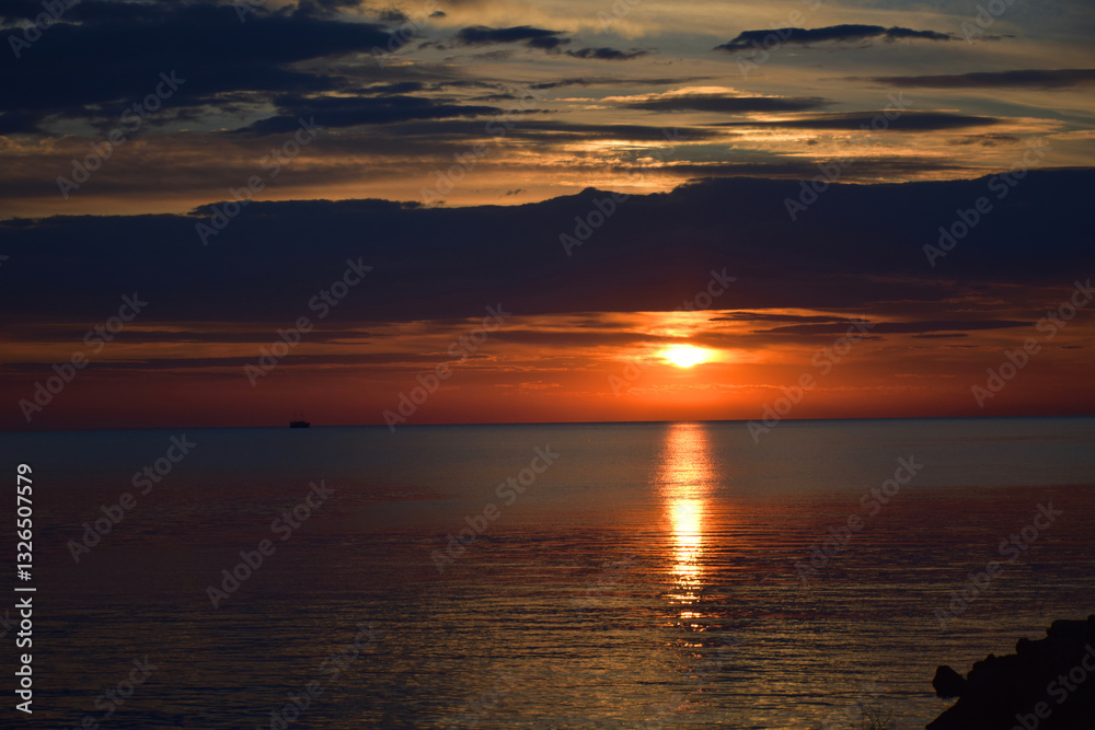 Naklejka premium Sunset Over the Ocean, Sea with Colorful Orange Sky, Reflections Rocky Shoreline. Sky with warm colors as the golden hour casts a stunning reflection on the sea water. Vacation spot for relaxation.