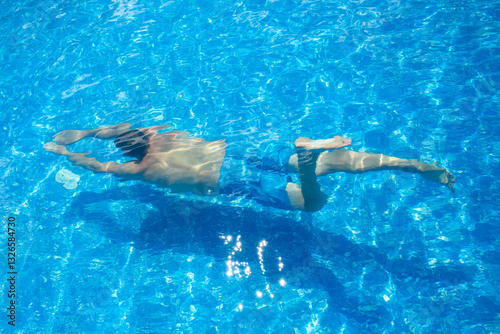 A young man in blue swimming trunks swims underwater in a pool with a blue bottom. The guy dived
