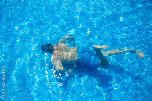 A young man in blue swimming trunks swims underwater in a pool with a blue bottom. The guy dived