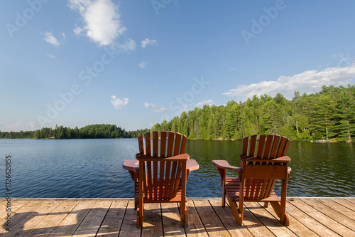 The calm blue lake in Muskoka reflects the summer sky, surrounded by lush trees. Two Adirondack chairs sit on a wooden dock, facing the water.