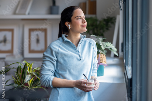 Beautiful sporty woman eating yogurt while listening music with earphone standing near the window at home. photo