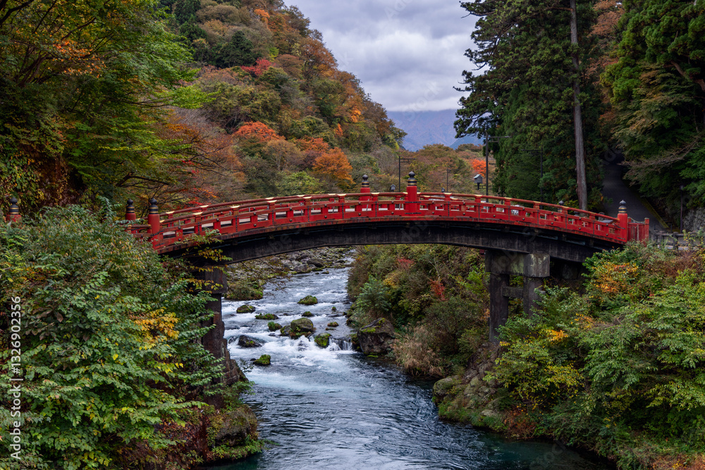 Historic red Shinkyo Bridge in Nikko Japan set against a forested hillside with vibrant autumn and Daiya River under an overcast sky in a scenic landscape Historic red Shinkyo Bridge in Nikko Japan set against a forested hillside with vibrant autumn and Daiya River under an overcast sky in a scenic landscape