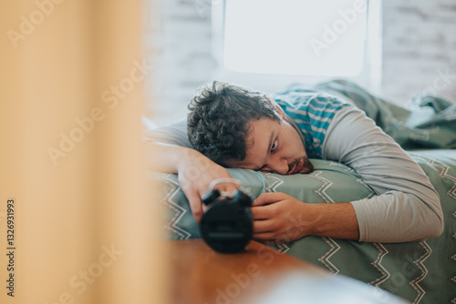 A tired man lies on a bed, holding a remote control, looking exhausted in the morning light. The image captures a feeling of fatigue and the struggle to get up.