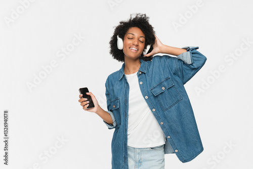 Happy young Afro American woman carried away with music dances carefree with arms raised keeps eyes closed wears stereo headphones on ears dressed in casual isolated over yellow background photo