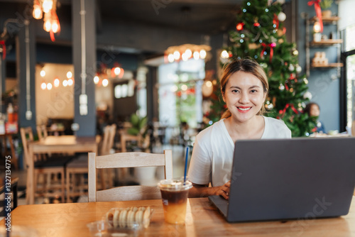 Asian woman in casual white shirt  working remotely on laptop in cafe  The background festive atmosphere with decorative hanging lights and Christmas tree. Blend of work and lifestyle.