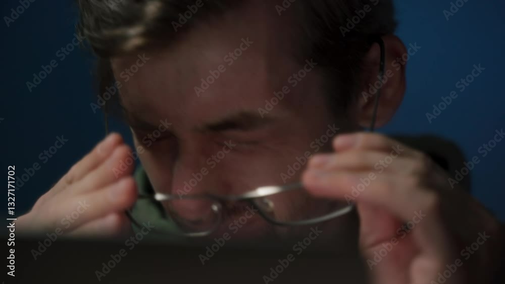 A tired coder wearing eyeglasses working late at night feels a headache and rubs his eyes from fatigue. Overwork concept