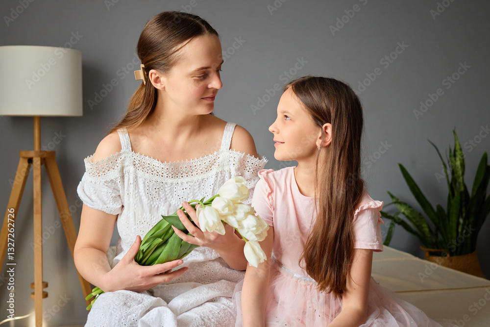 Obraz premium A young mother and her daughter sit on a bed in a cozy room, sharing a joyful moment. The mother holds a bouquet of white flowers, and they exchange warm gazes