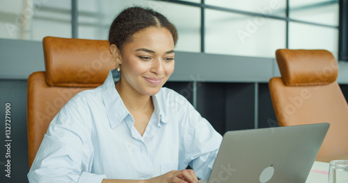smiling young woman working on a laptop in a modern office setting, sitting in a leather chair and focusing on her screen with a happy expression