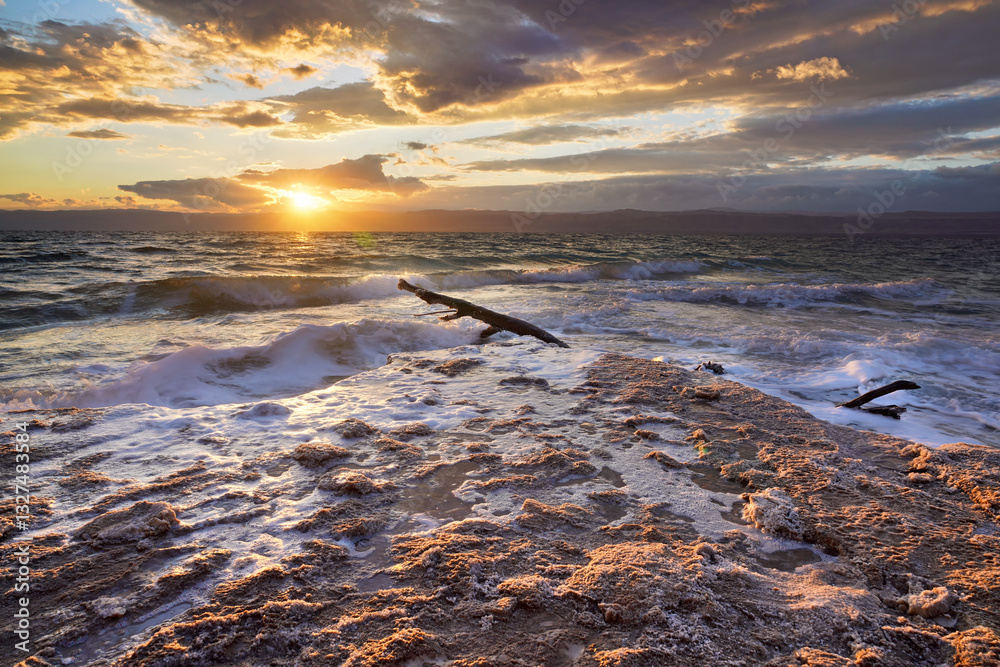 Sun shines through dramatic sunset clouds to waves at Jordanian side of Dead Sea, shore sand covered with salt