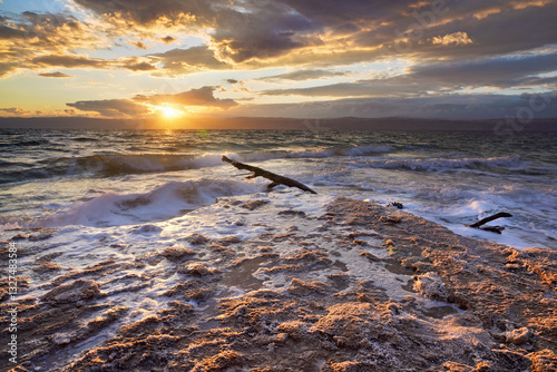 Obraz na płótnie Sun shines through dramatic sunset clouds to waves at Jordanian side of Dead Sea, shore sand covered with salt w salonie