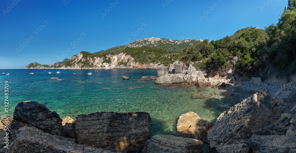 Calm clear green sea near shore with large rocks, sun shines to small waves, boats at distance, typical scenery at one of many bays in Liapades, Greece