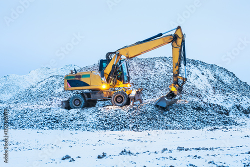 A excavator on a winter morning at the construction site, standing on a pile of dirt covered with snow