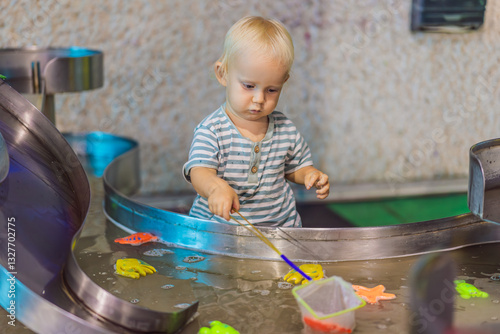 Toddler boy catching toy fish with a net in a vibrant water play area at an entertainment center. A joyful moment of learning, play, and discovery designed to inspire young children