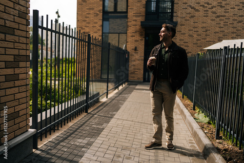 Full length portrait of handsome young man holding coffee cup standing by residential building with metal fence and brick walls, smiling looking away, enjoying peaceful moment outdoors.