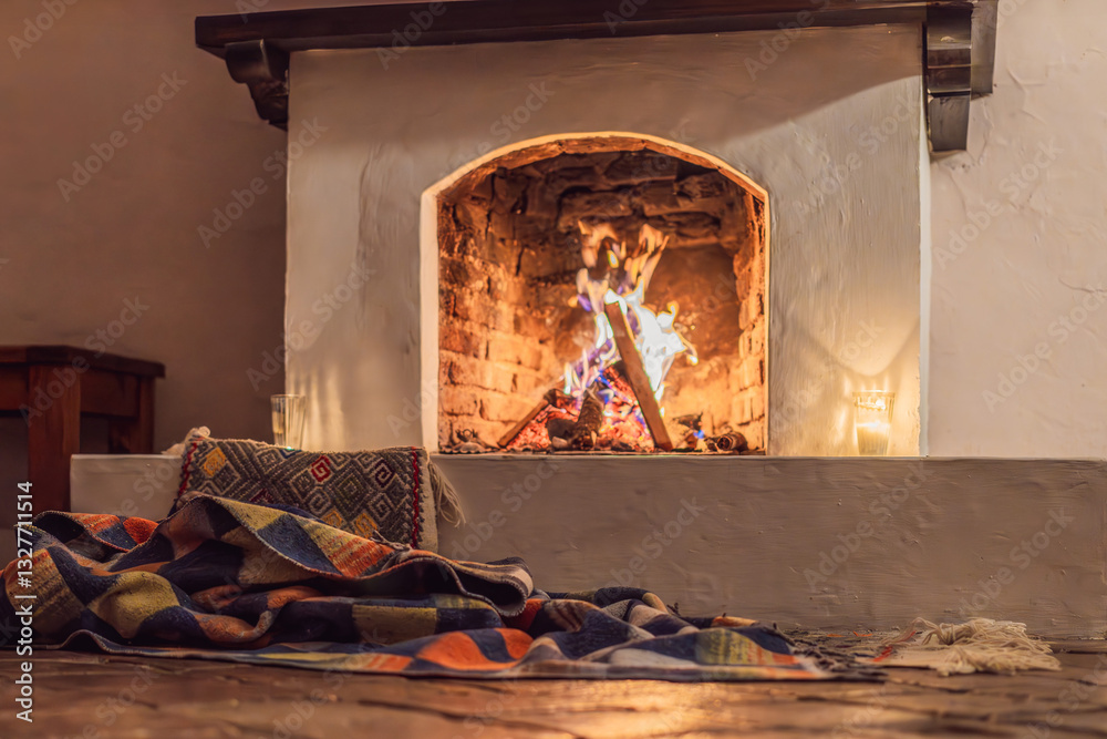 A warm and inviting scene featuring a lit fireplace with glowing firewood and candles. A colorful traditional rug and pillow lie on the floor, creating a cozy atmosphere. Home, relaxation, and rustic