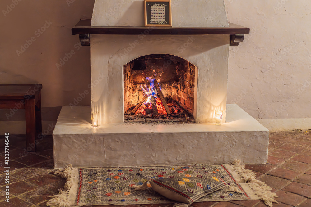 A warm and inviting scene featuring a lit fireplace with glowing firewood and candles. A colorful traditional rug and pillow lie on the floor, creating a cozy atmosphere. Home, relaxation, and rustic