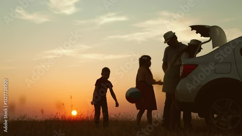 Playful family at sunset by car. cheerful children parents going on travel road trip by automobile packing baggage putting luggage in trunk kids playing ball. Packing preparation for family picnic.