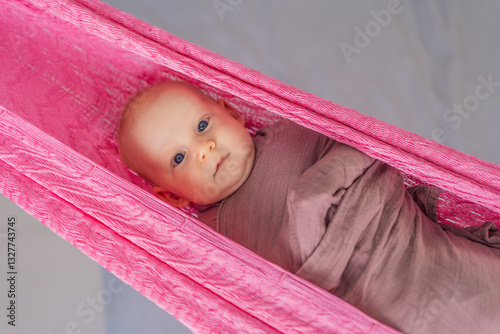 A 3-month-old baby peacefully falls asleep in a soft muslin swaddle while cradled in a cozy hammock. A serene and tender moment of comfort and relaxation. Baby care, rest, and tranquility concept