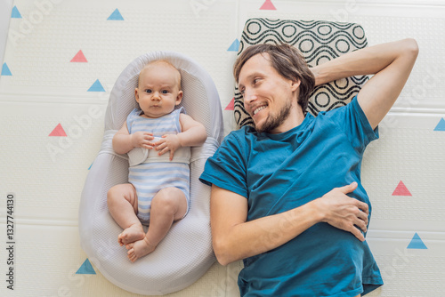 Obraz na płótnie A loving father lies next to his 3-month-old baby, safely secured in a cozy infant cocoon with a special safety belt. Heartwarming family bonding moment. Parenthood, love, and baby care concept w salonie